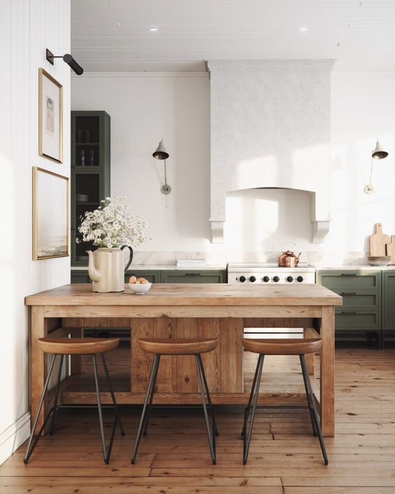 a chic modern farmhouse kitchen with green cabinets, a large white hood, a small dining zone with stained furniture