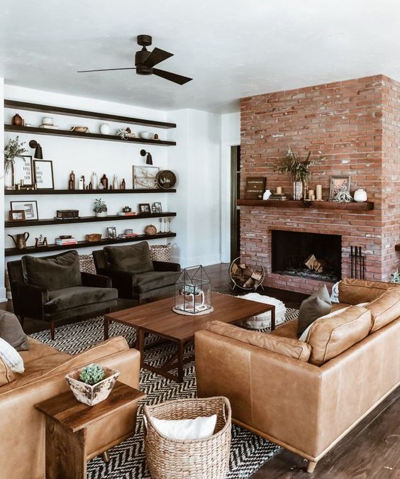 a chic modern farmhouse living room with a red brick fireplace, dark chairs, a tan leather sofa and chair, a coffee table and dark-stained shelves