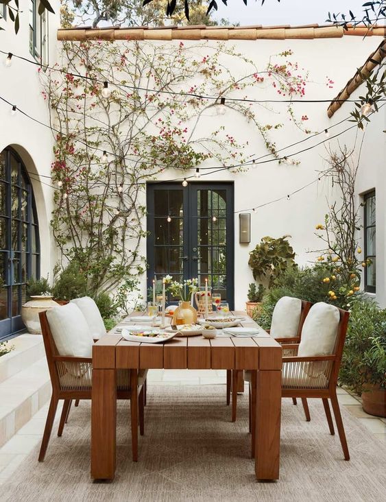 a chic modern farmhouse terrace with a stained table and chairs, greenery and blooms and string lights over the space