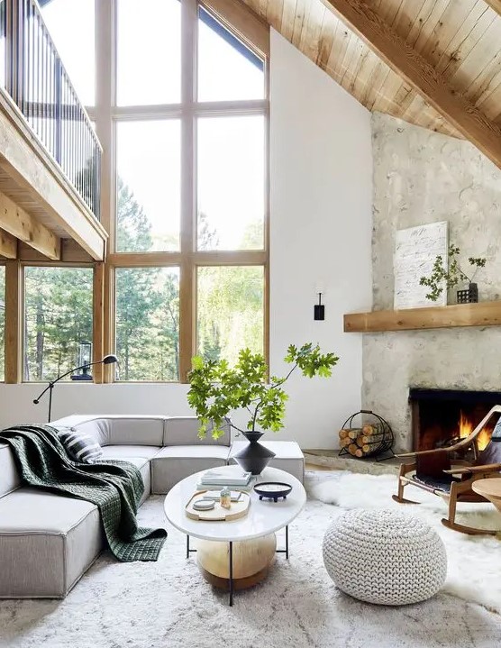 a contemporary living room with a fireplace clad with stone, a grey low sectional, an oval table, a crochet pouf, greenery branches and a floor lamp