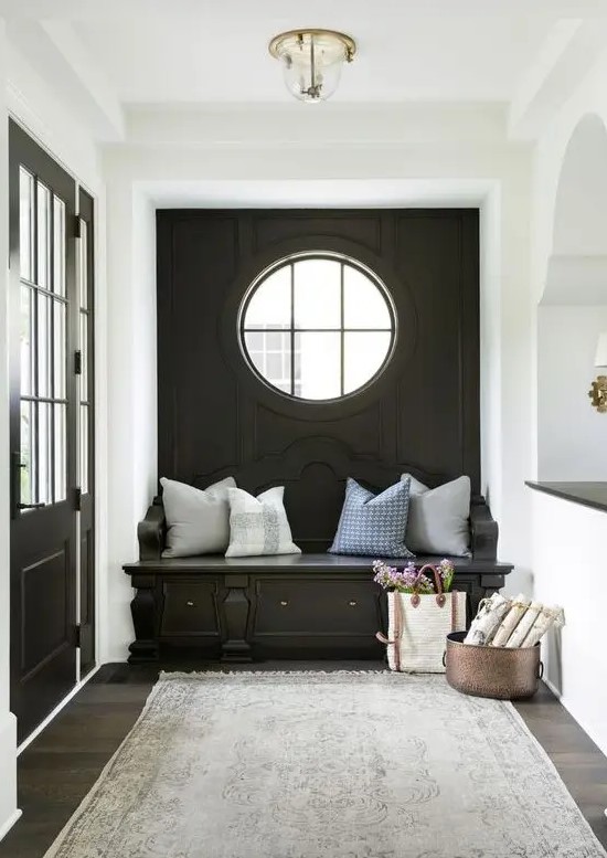 A cozy black and white mudroom with a black accent wall, a porthole window and a built in bench, neutral pillows and a grey rug
