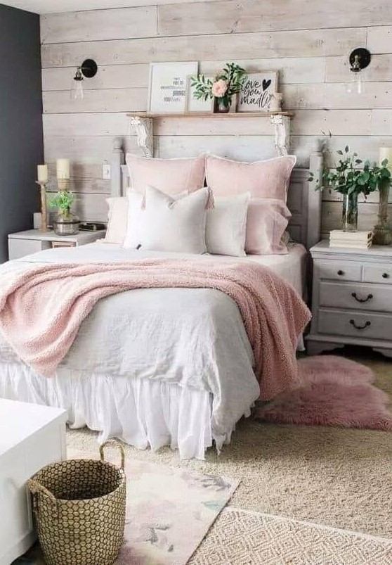 a cozy farmhouse bedroom with a whitewashed wooden wall and whitewashed furniture, pink bedding, greenery and a basket for storage