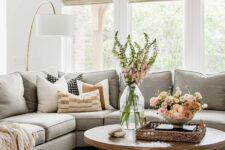 a cozy modern farmhouse living room with a grey sectional, a low coffee table, amber leaher poufs, some blooms and printed pillows