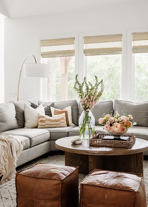 a cozy modern farmhouse living room with a grey sectional, a low coffee table, amber leaher poufs, some blooms and printed pillows
