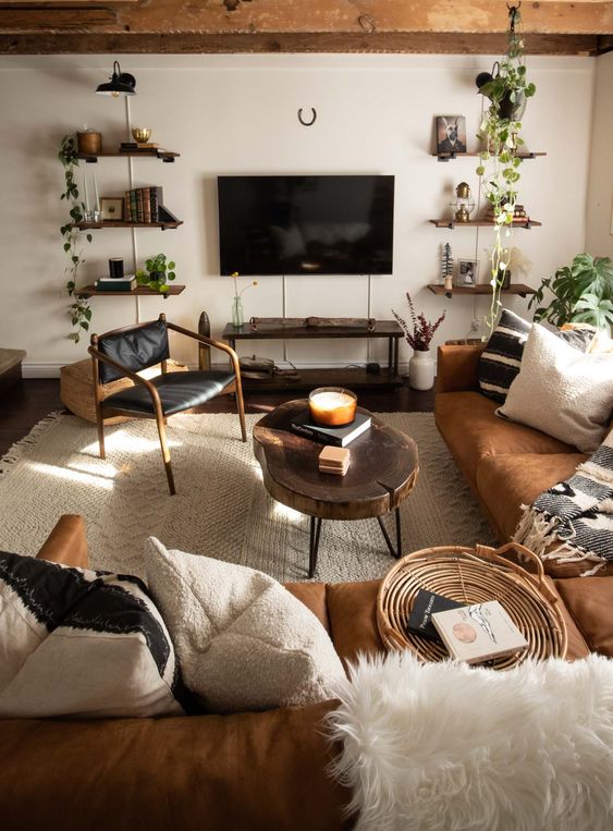 a cozy modern farmhouse living room with wooden beams and wooden shelves, an amber leather sectional, a black leather chair and some greenery