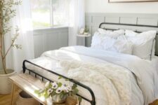 a cute and welcoming modern farmhouse bedroom with grey paneled walls, a wrought bed with neutral bedding, a stained bench and baskets, a potted tree