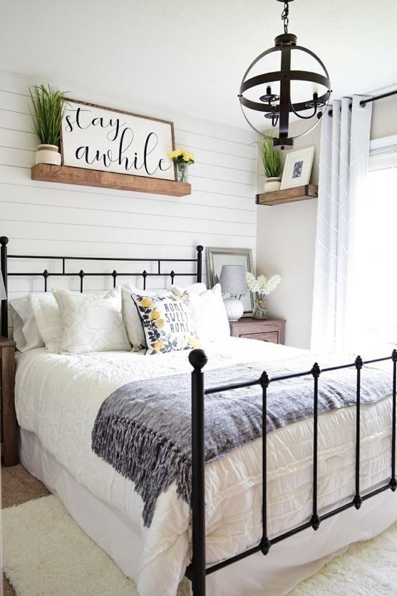 a cute modern farmhouse bedroom with white wooden plank walls, a forged bed, a sphere chandelier, shelves and greenery