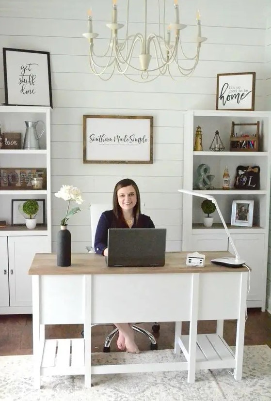 A cute white modern country home office with a planked wall, built in storage units, a large desk and a neutral chandelier