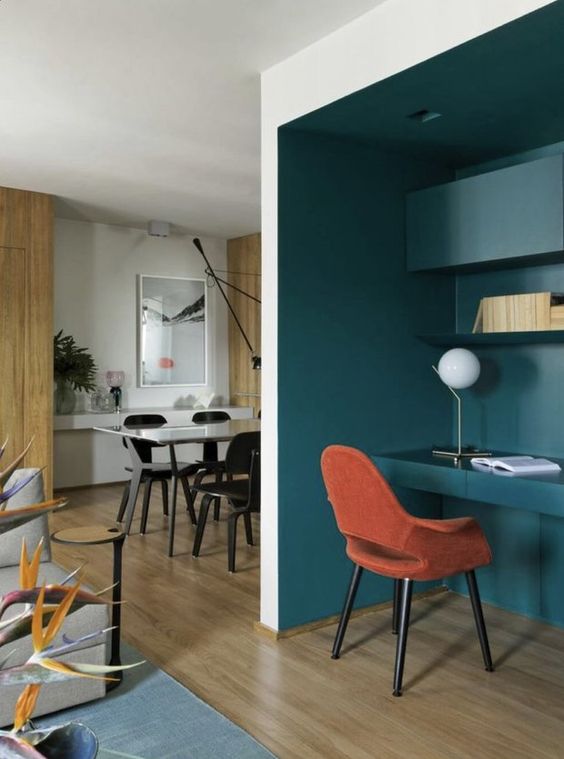 A deep teal niche with built in shelves and a desk, an orange chair, a table lamp and built in lights is great for working
