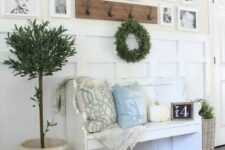 a farmhouse entryway with a white bench with pillows, a pumpkin, a gallery wall, a greenery wreath and a potted tree