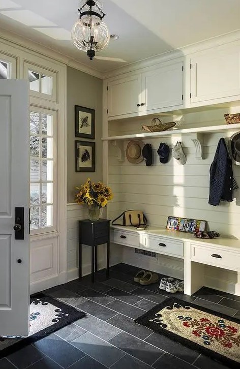 A farmhouse entryway with an ivory wooden furniture piece iwth a built in bench of drawers and more storage space plus dark tiles on the floor for a contrast
