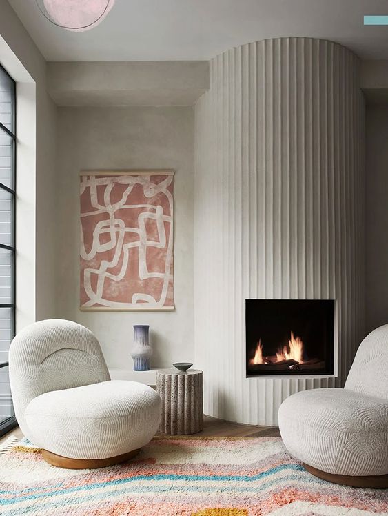 a fireplace with a fluted surround, neutral curved chairs, a bold printed rug and a bold artwork look so chic together