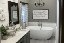 a grey farmhouse bathroom with a large vintage vanity, a stone countertop, an oval tub, a chandelier and some vintage wall lamps