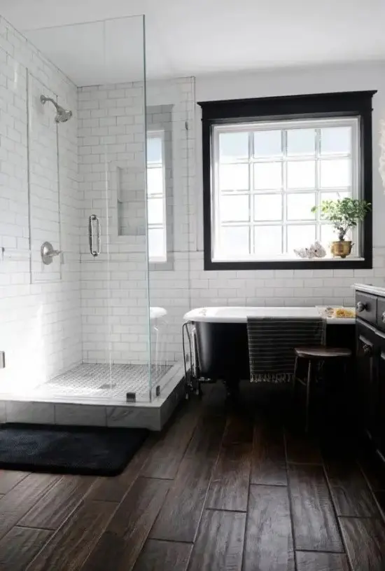 a laconic monochromatic farmhouse bathroom with a dark wooden floor, white subway tiles, a black clawfoot bathtub and a window