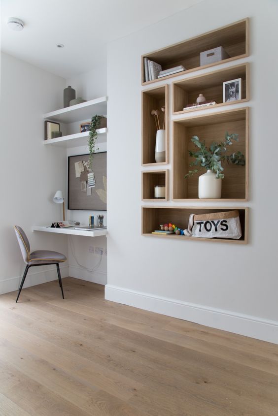 A large wall niche with built in shelves and a desk, potted greenery, a memo board, some books and vases and a grey chair