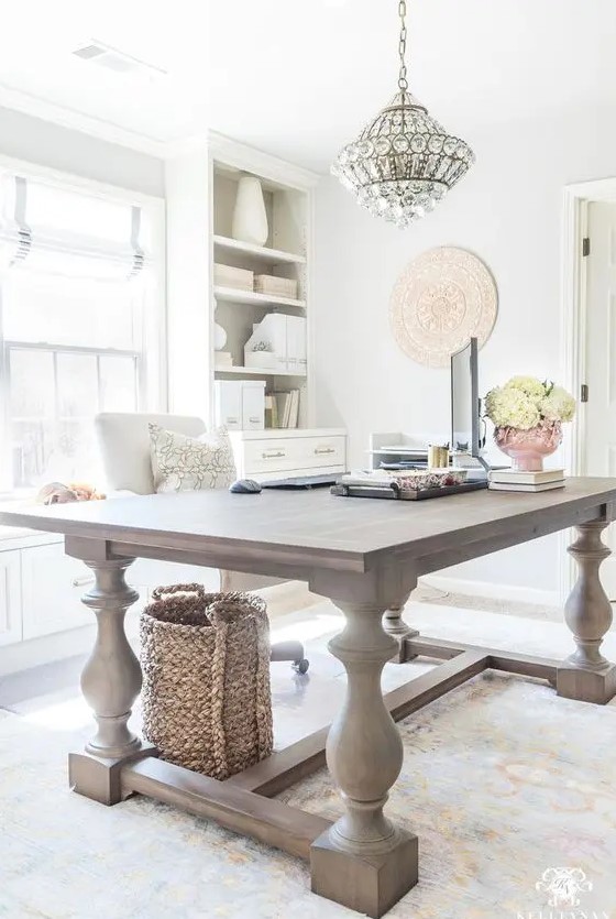 a light-filled farmhouse home office with a large wooden desk, neutral storage units, a crystal chandelier and neutral blooms