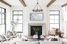 a light-filled modern farmhouse living room with reclaimed beams, a fireplace, neutral seating furniture, greenery and a heavy chandelier