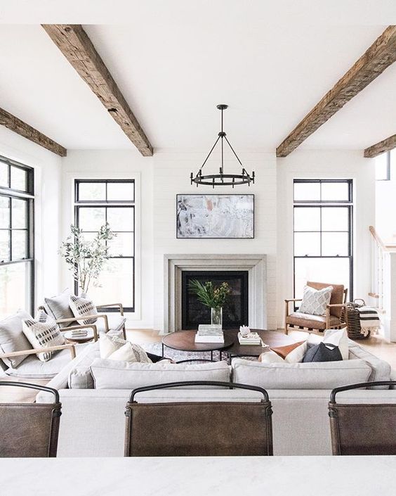 a light-filled modern farmhouse living room with reclaimed beams, a fireplace, neutral seating furniture, greenery and a heavy chandelier