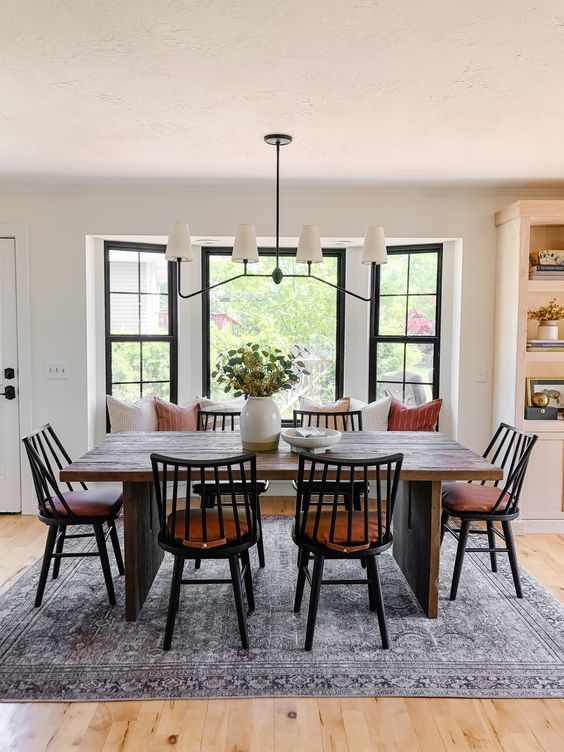 a lovely modern farmhouse dining nook with a windowsill seat, a stained table, black chairs, a chandelier and a printed rug