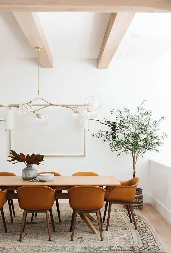 a lovely modern farmhouse dining space with wooden beams on the ceiling, a wooden table, amber chairs, a large and chic chandelier and a potted tree