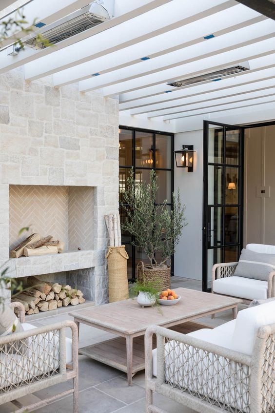 a lovely modern farmhouse terrace with a fireplace clad with stone, a low whitewashed coffee table, neutral wicker chairs and potted greenery