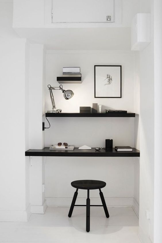 a minimalist niche with a built-in shelf and desk, a table lamp, some laconic decor and a black stool