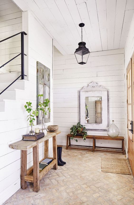 a modern and chic farmhouse entryway with a large mirror, a bench and a console table, a brick floor and some greenery