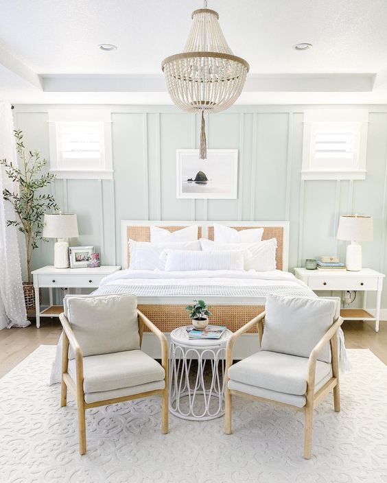 a modern coastal farmhouse bedroom with pale blue paneled walls, a cane bed, pale blue chairs, white nightstands and a bead chandelier