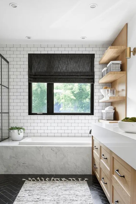 a modern country bathroom with white subway tiles and black herringbone ones, a bathtub clad with white marble, light stained furniture and a window with a shade