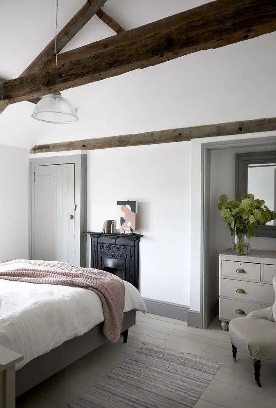 A modern country bedroom with dark stained wooden beams, a non workign fireplace, neutral furniture and neutral textiles