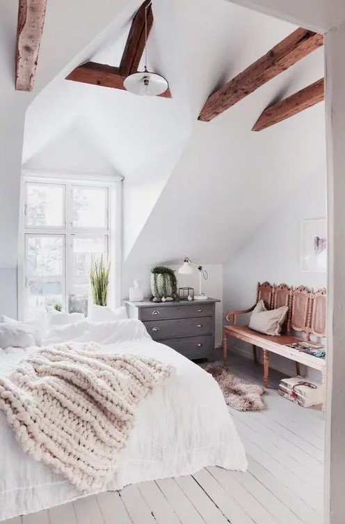 a modern country bedroom with stained beams on the ceiling, a carved wooden bench and a bed with neutral bedding, a potted plant and some decor