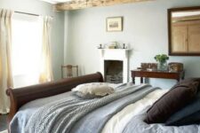 a modern country bedroom with wooden beams, a fireplace, a dark stained bed and a mirror plus some vintage furniture