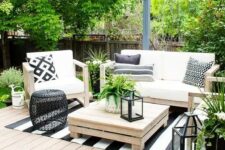 a modern country terrace with a deck, simple wooden furniture and printed textiles, a black side table and candle lanterns and greenery
