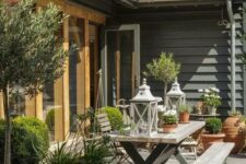 a modern country terrace with a wooden dining set including benches, potted greenery, candle lanterns and potted trees is cool