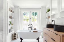 a modern farmhouse bathroom with a pebble floor, shiplap, a timber vanity, black sinks and a clawfoot tub by the window