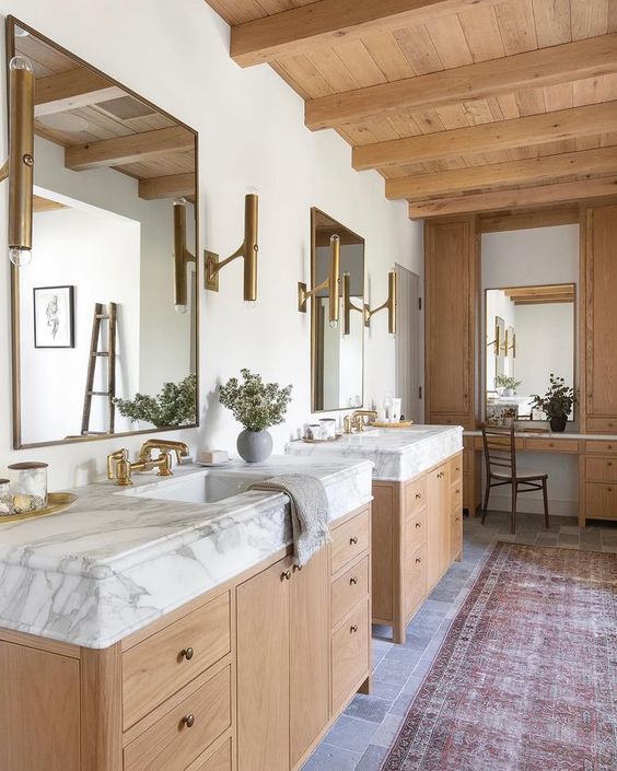 a modern farmhouse bathroom with a wooden ceiling with beams, wooden cabinetry and vanitites, mirrors and gold sconces