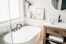 a modern farmhouse bathroom with grey and white tiles, a stained vanity, an oval tub, a shower space, a round mirror and some decor