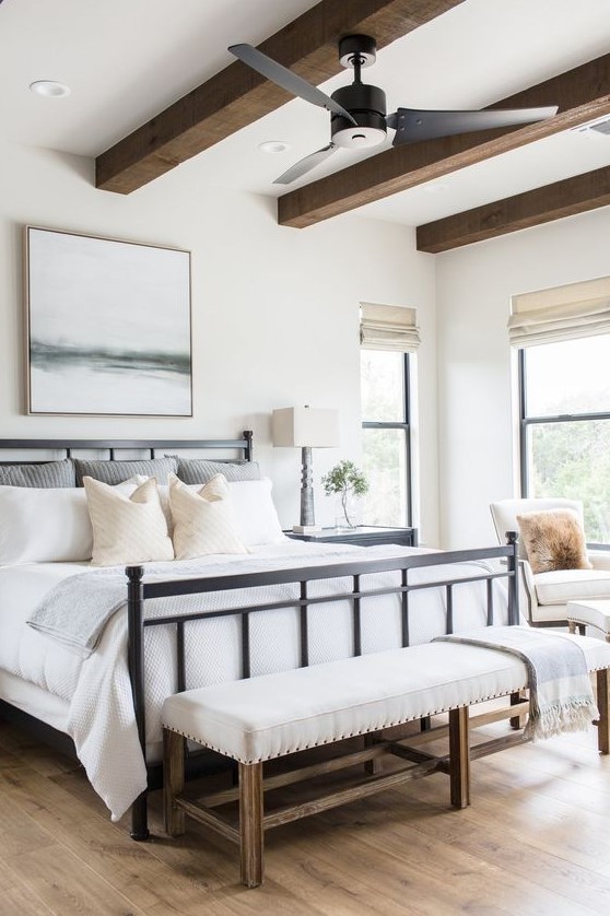 a modern farmhouse bedroom with a black forged bed, a neutral upholstered bench, wooden beams and neutral chairs