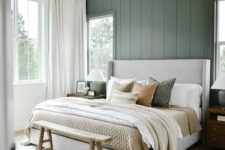 a modern farmhouse bedroom with a green accent wall, a grey upholstered bed with neutral bedding, a wooden bench and a chandelier