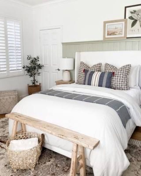 a modern farmhouse bedroom with a green shiplap wall, a white upholstered bed with printed bedding, a stained bench, some art and baskets