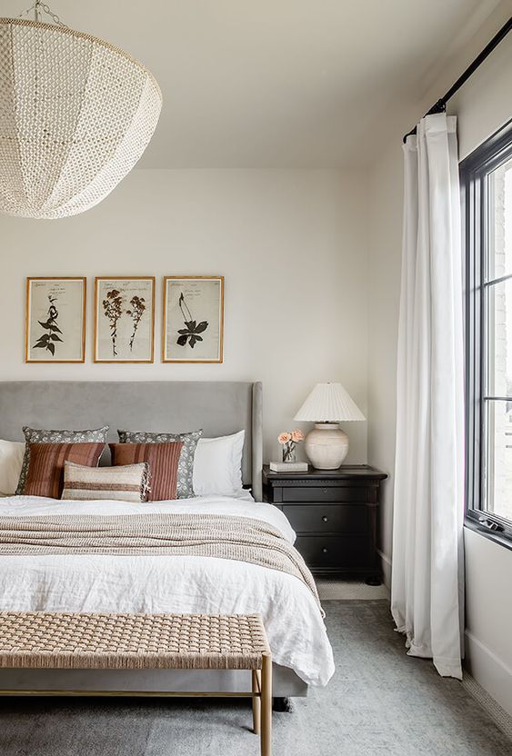 a modern farmhouse bedroom with a grey upholstered bed and neutral bedding, a woven bench, black nightstands and artwork