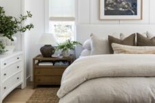 a modern farmhouse bedroom with a white upholstered bed with neutral bedding, a wooden bench, stained nightstands, a white dresser and decor