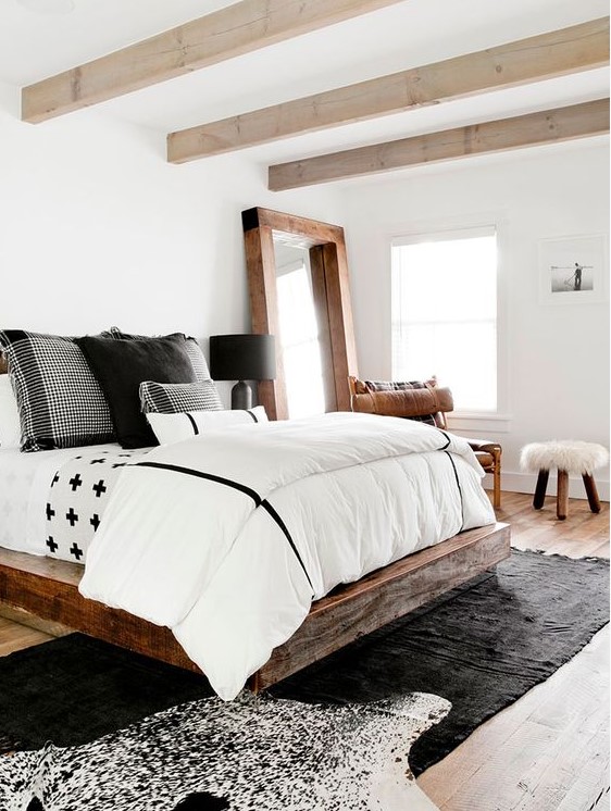 a modern farmhouse bedroom with light-stained wooden beams, a rich-stained wooden bed, black and white bedding and layered rugs