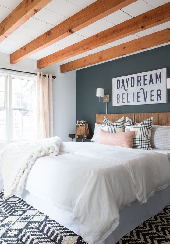 a modern farmhouse bedroom with rich-stained wooden beams on the ceiling, a stained bed, neutral bedding, a black accent wall and a printed rug
