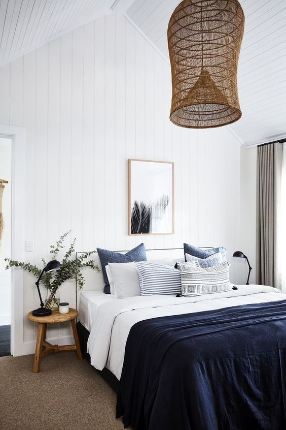 a modern farmhouse bedroom with shiplap walls, a white bed with navy and white bedding, stained stools and a woven pendant lamp