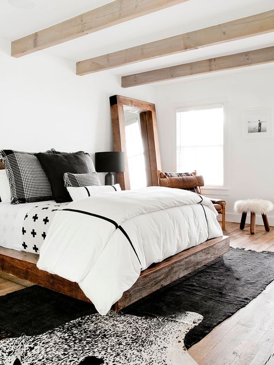 a modern farmhouse bedroom with wooden beams, a dark-stained bed with contrasting bedding, a leather chair, layered rugs and a large mirror