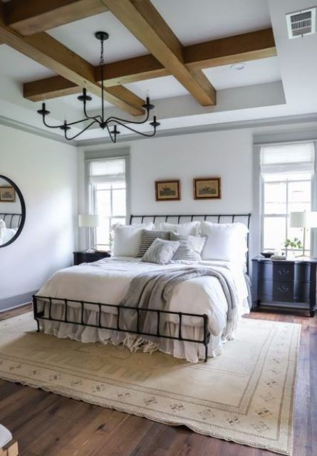 a modern farmhouse bedroom with wooden beams, a wrought ceiling, black nightstands, neutral bedding and a chandelier