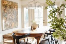 a modern farmhouse dining room with a rounded stained table, black chairs, a bench, a large scale artwork and a cool pendant lamp