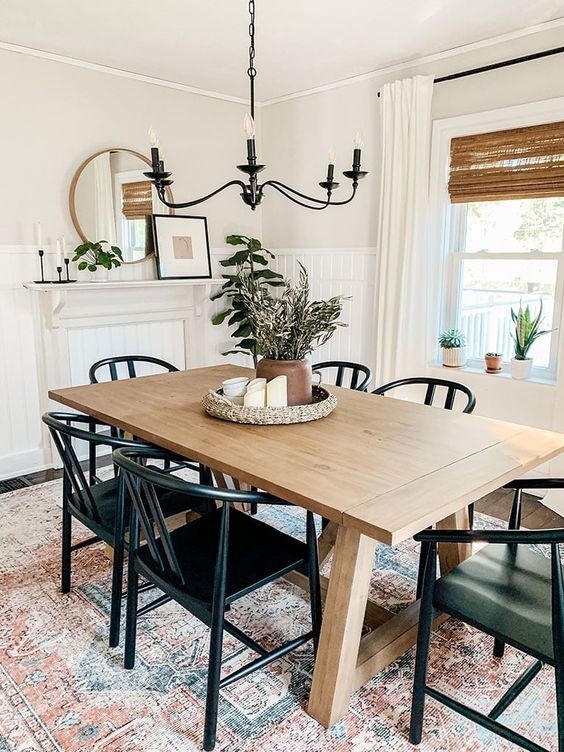 a modern farmhouse dining space with a faux fireplace, a stained table, black chairs, a black chandelier and some potted plants
