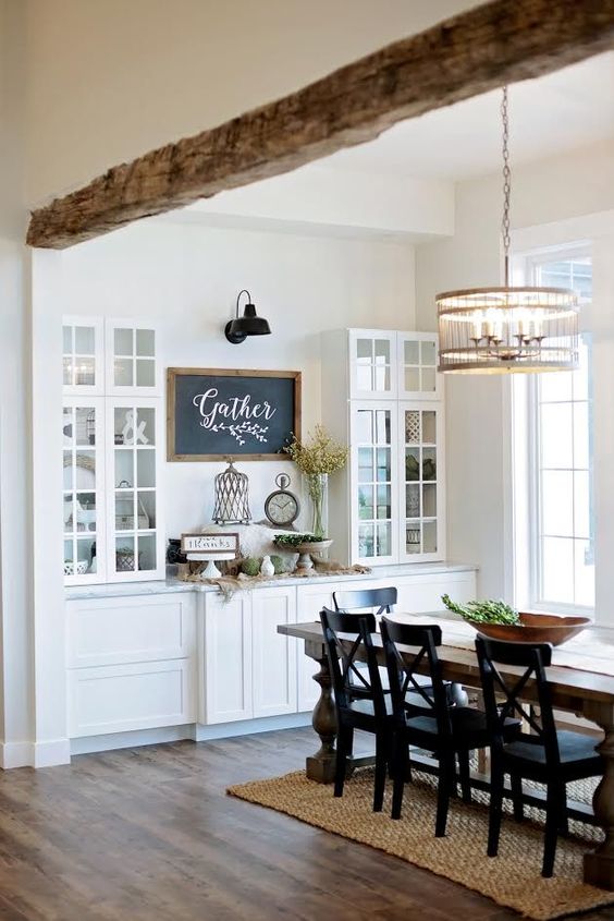 a modern farmhouse dining space with a long stained table, black chairs, built-in buffets and a cabinets, a wooden beam and a chandelier